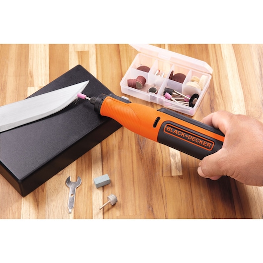 Photo of BLACK+DECKER BCRT8K35 rotary tool in use sharpening a knife, accessories case visible on wooden surface.