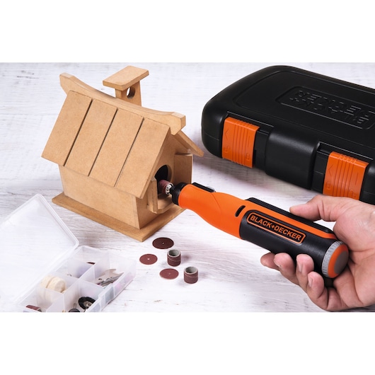 BLACK+DECKER BCRT8K35 rotary tool sanding a wooden birdhouse, with accessories and carrying case on white surface.