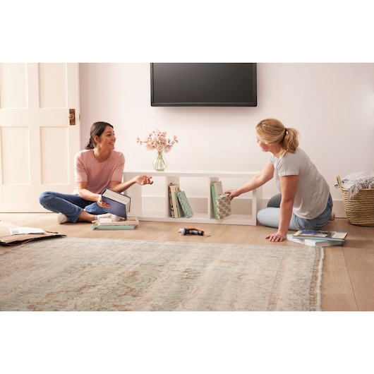 Photo of BLACKDECKER BCRTA601I cordless screwdriver on floor between two women assembling a white bookshelf in a living room.