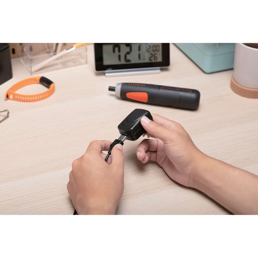 Photo of BLACKDECKER BD40K27 electric screwdriver on desk with hands plugging in charger, orange wristband nearby.