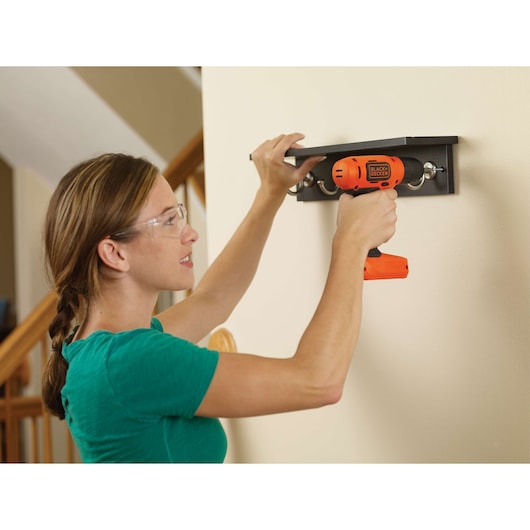 Photo of person using BLACKDECKER BDCD8C orange cordless drill to install a wall-mounted shelf with hooks indoors.