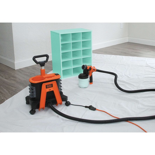 A BLACK+DECKER paint sprayer system (BDPH1200) in orange and black is shown placed on a white sheet in a room with wooden floors and white walls. The paint sprayer is connected to a hose, and there is a small, mint green shelving unit nearby.