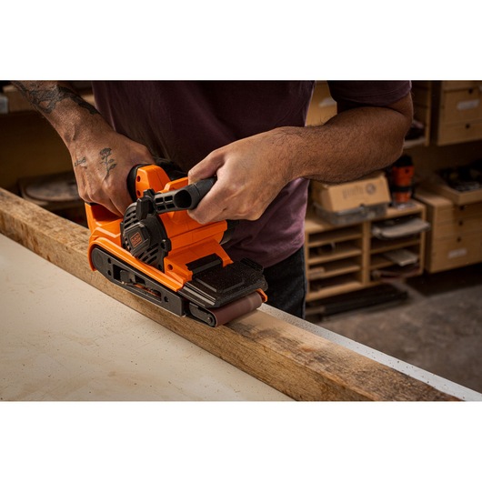 Photo of BLACKDECKER belt sander BEBS0900 smoothing wood plank in workshop setting.
