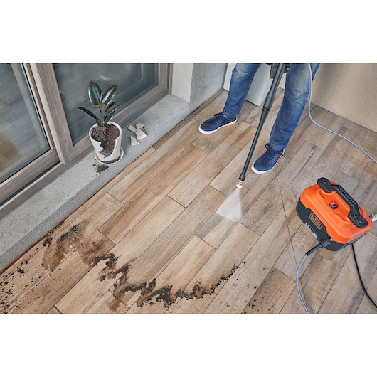 BLACKDECKER BEPW1600 pressure washer cleaning dirt from patio tiles, orange unit and water spray visible, top view.