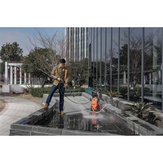 Person using BLACKDECKER BEPWB2400 pressure washer to clean outdoor stone surface near building with plants.
