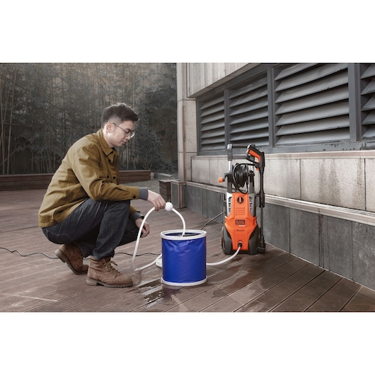 BLACKDECKER BEPWB2400 pressure washer shown outdoors with hose in blue water bucket and a person kneeling nearby.