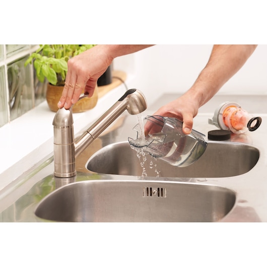 BLACKDECKER BHFEA18D1 vacuum dust container being rinsed under kitchen faucet in stainless steel sink.