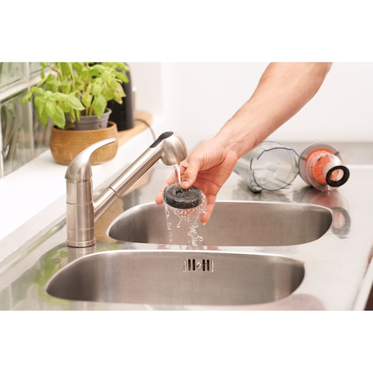 Photo of BLACKDECKER BHFEA18D1 vacuum filter being rinsed under a kitchen tap, vacuum dust container on sink.