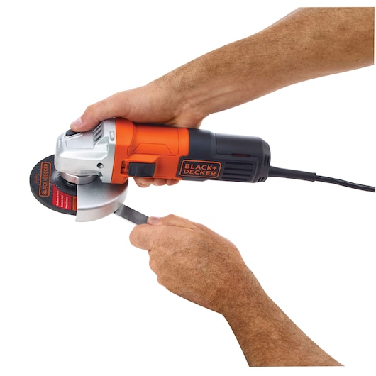 Photo of BLACK+DECKER G650 angle grinder in use, orange and black body, hands holding tool and metal piece, side view.