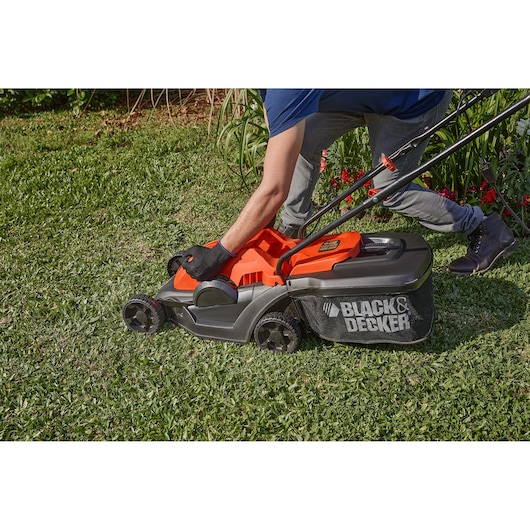 Photo of BLACK+DECKER GR3400 lawn mower in use on grass, side view, orange and black design, mesh grass bag visible.