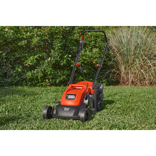 A BLACK+DECKER electric lawn mower with a bright orange and black design is shown on a grassy lawn with green shrubs in the background.
