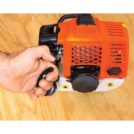 BLACKDECKER GST26 orange and black engine close-up with hand pulling starter cord, top view.