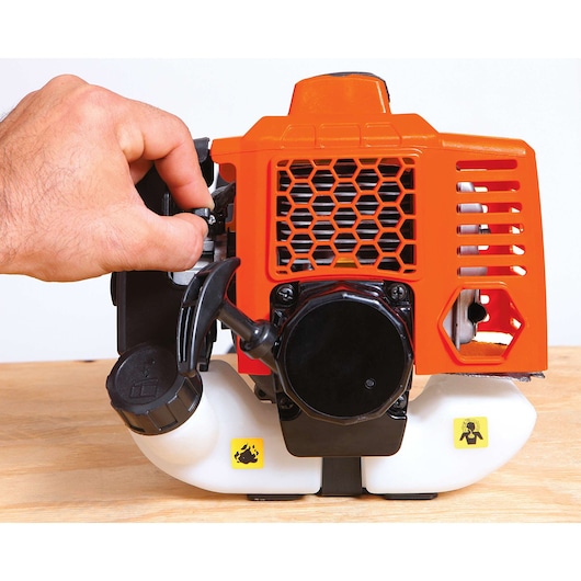 BLACKDECKER GST26 gas trimmer engine close-up, orange housing, fuel tank visible, hand operating control, side angle.