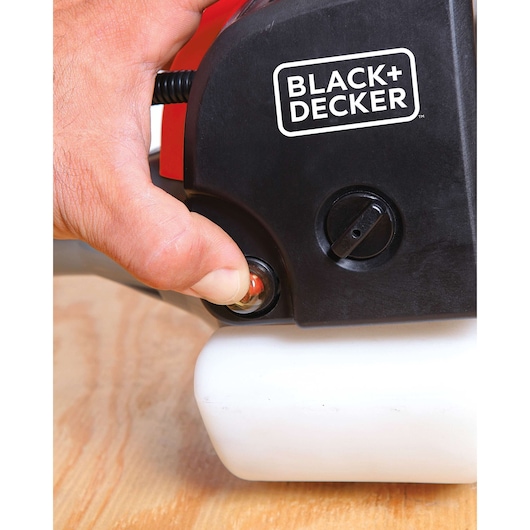 Close-up of hand pressing primer bulb on BLACK+DECKER GST26, black and white trimmer body, wooden surface.