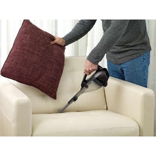 BLACKDECKER HLVA320J00 handheld vacuum cleaning cream sofa under red pillow indoors.