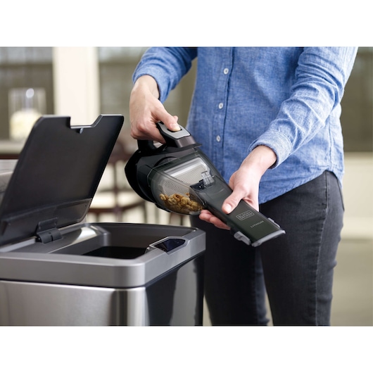 BLACKDECKER HLVA320J00 handheld vacuum being emptied into a trash can, close-up side view.