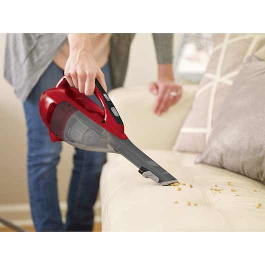 BLACKDECKER HLVA320J26 handheld vacuum in red being used to clean crumbs from a beige sofa.
