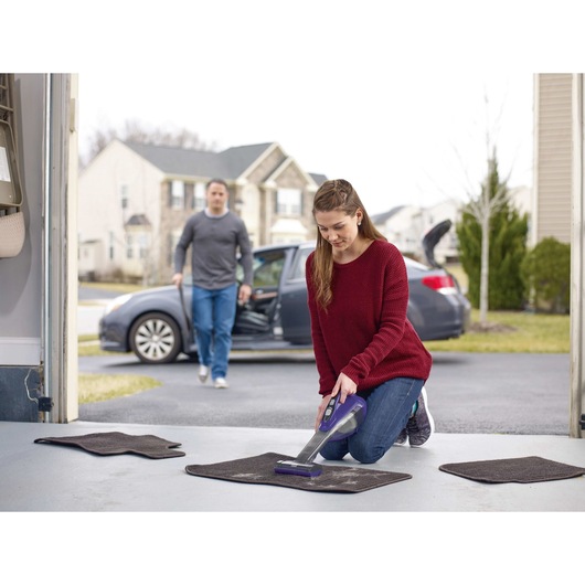 BLACKDECKER HLVA325JP07 handheld vacuum cleaning car mats in driveway, person kneeling and using device.