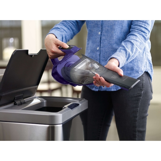 Person emptying BLACKDECKER HLVA325JP07 handheld vacuum dustbin into a trash can, indoor setting.