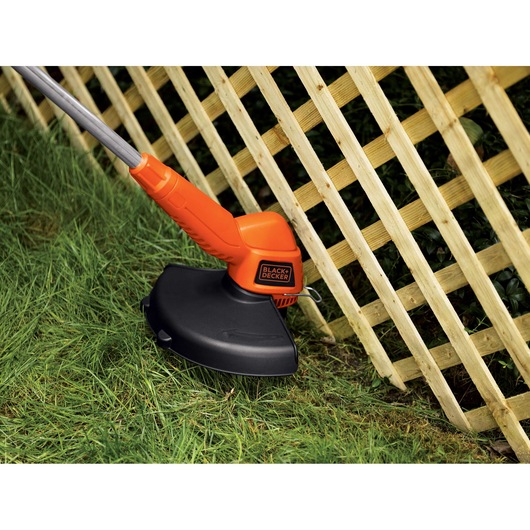 BLACKDECKER ST7700 orange and black grass trimmer cutting lawn near a wooden lattice fence.