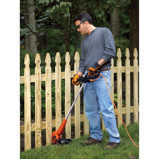 Photo of a person using a BLACKDECKER ST7700 orange corded string trimmer near a wooden fence in a yard.