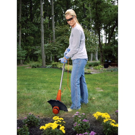 Person using BLACKDECKER ST7700 corded string trimmer on lawn near flower bed in garden setting.